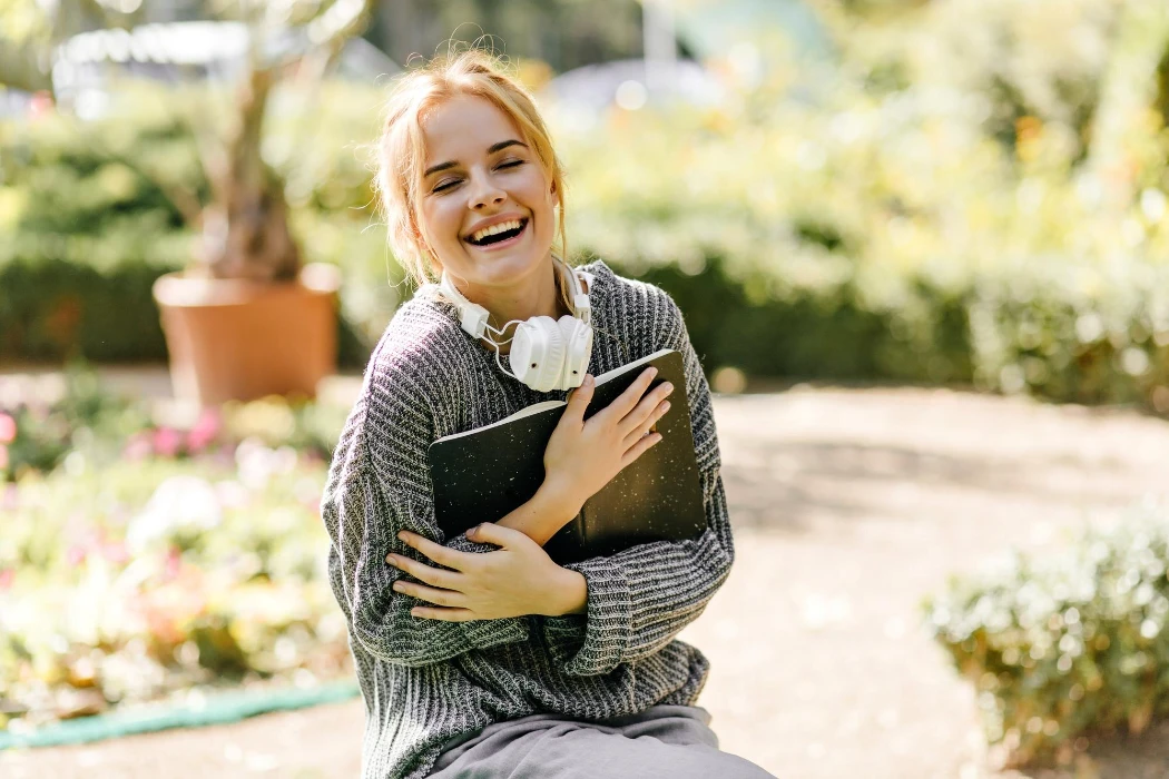 Happy caucasian girl with headphones laughing in autumn garden Blonde female student posing with book and smiling